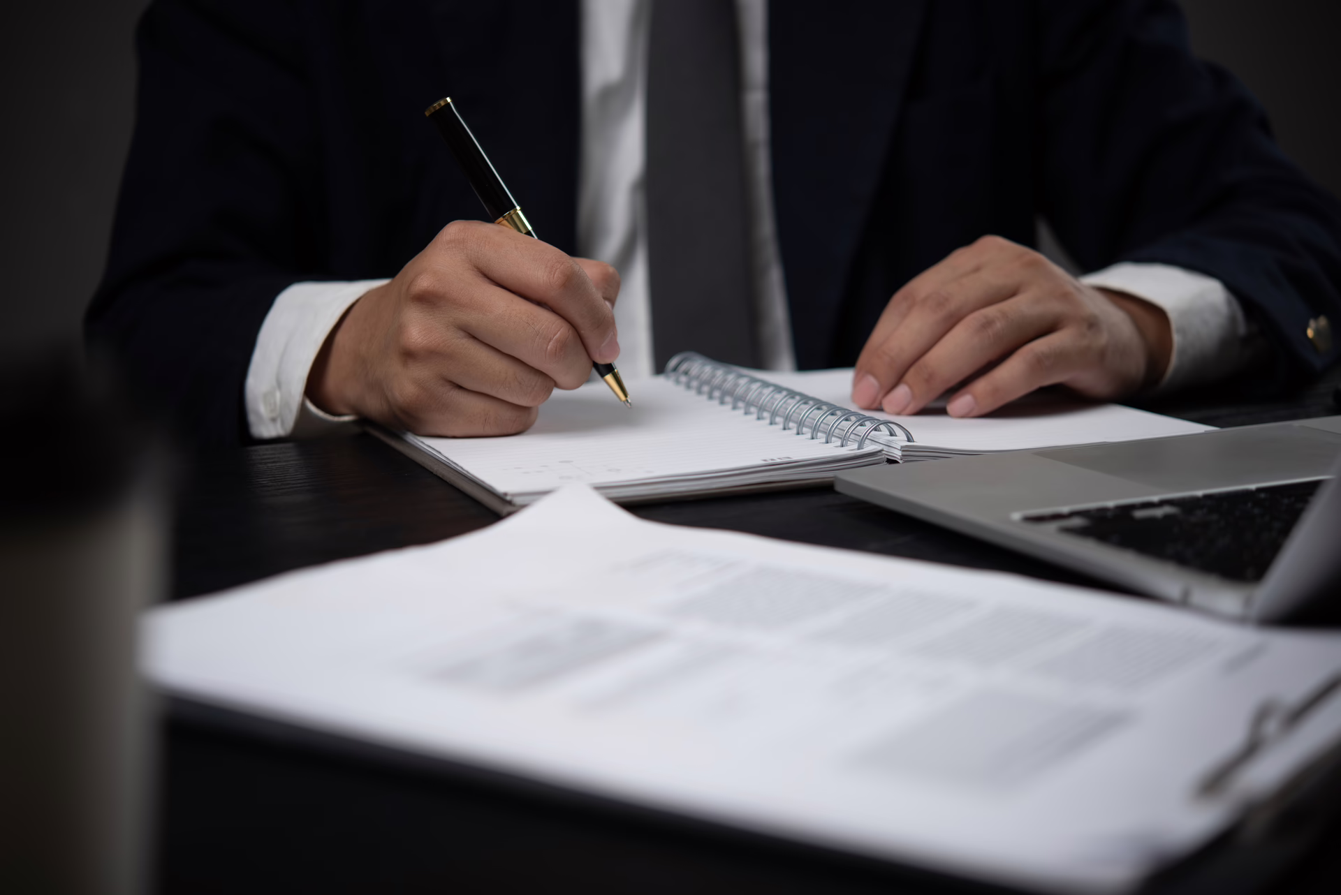 Person making notes in a notebook at a desk with legal documents and a laptop in a professional office setting.
